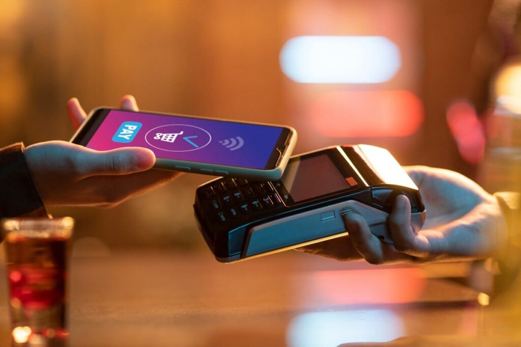 Una persona sostiene un teléfono móvil con la pantalla mostrando una aplicación de pago digital, acercándolo a una terminal de punto de venta para realizar una transacción sin contacto en un ambiente con luces cálidas
